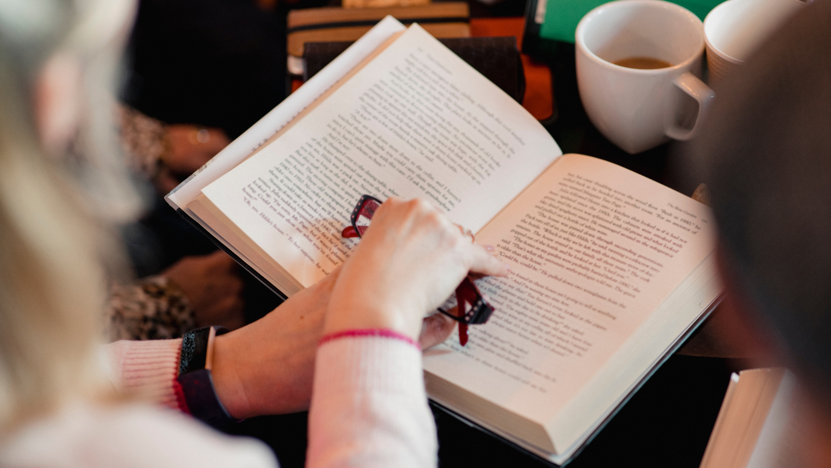 woman reads open books with friends while drinking coffee and holding reading glasses 