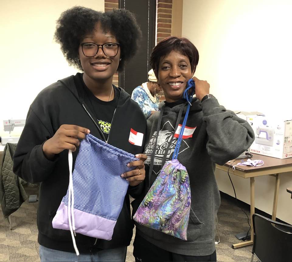Two women are smiling and showing us the drawstring bags they finished in the sewing class.