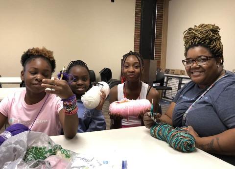 Photo of a group of adults and teens sitting at a table, crocheting.