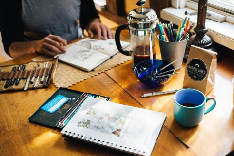 Teen artist with their artwork and tools out, drawing a picture at a table using natural sunlight.