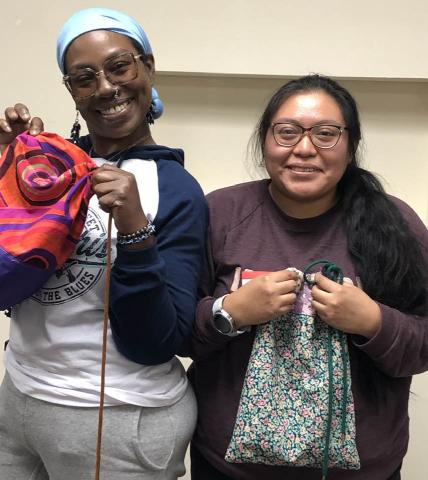 Two women are smiling and showing us the drawstring bags they finished in the sewing class.