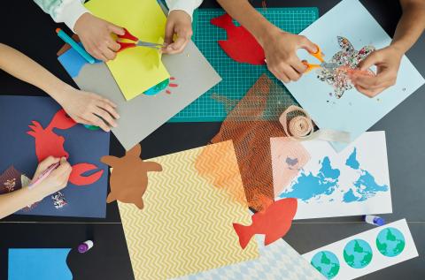Scene of children sitting at a table with colored-paper, scissors, glitter, 