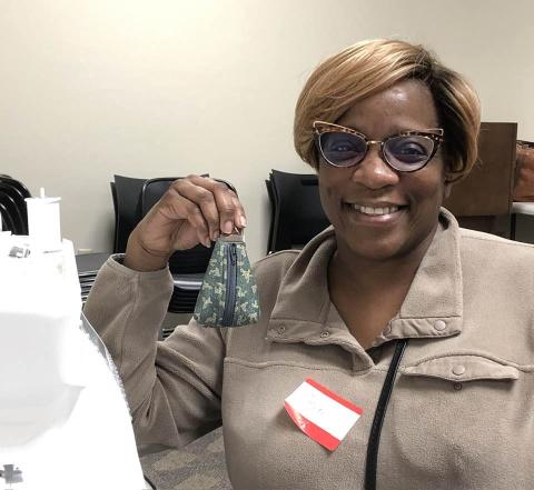 Person sitting at table in front of sewing machine, smiling and holding up her finished project, a small pouch with a zipper.