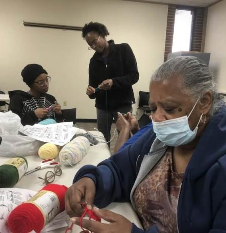 Three people sit at a table in the Cherokee meeting room, working on crochet projects.