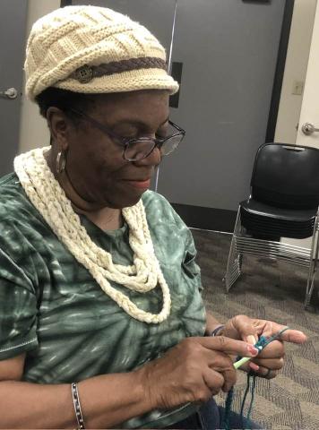 Woman in green tie-dye shirt, wearing a crocheted necklace and cap works on another project at the Cherokee Crochet Club.