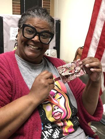 Woman in red sweater and black glasses has a huge smile as she holds up her finished sewing project at the Cherokee Library.