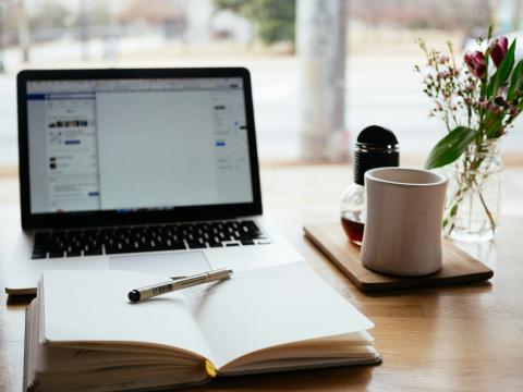 Laptop and notebook open, ready for creative writing. A cup of coffee is next to them on the table.