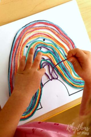 photo of a young child's hands placing yarn on a piece of paper. Other pieces of yarn have been glued down already to make the shape of a rainbow.