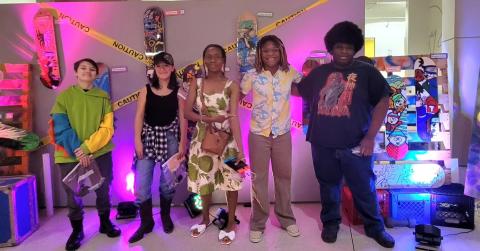 5 teens standing next to each other, posing by their custom designed skateboard.