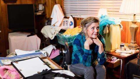 older women in blue shirt sits among clutter in her home