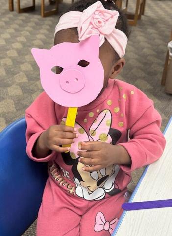 Small girl in pink outfit holds up a pink pig mask during the Cherokee Story Time.