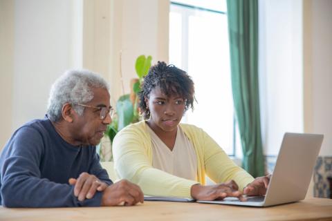 A senior and adult sit next to each other while using a computer.