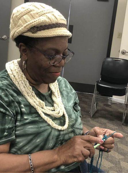 Lady in tie-dye shirt, crocheted hat and necklace is crocheting at the Cherokee Library.