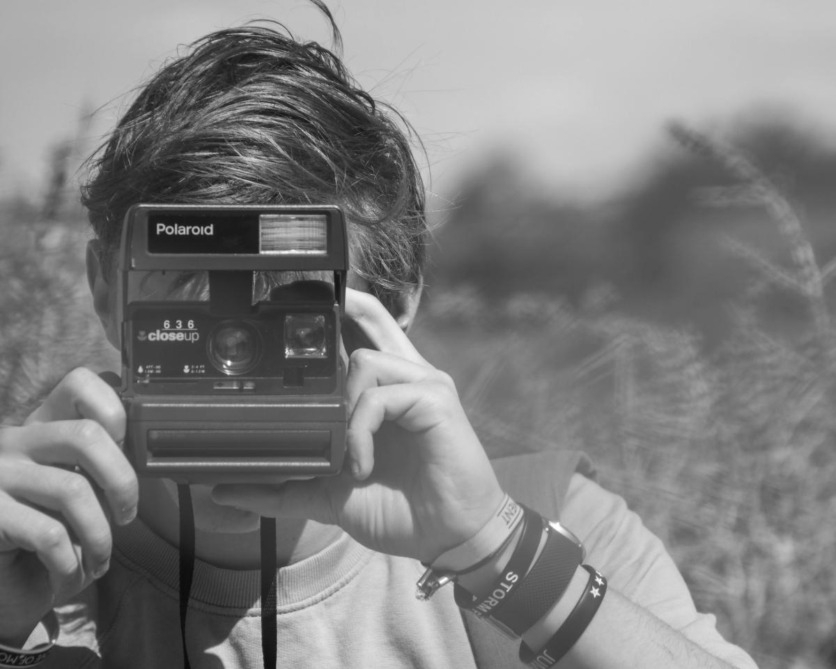 A young man holds a polaroid camera pointed at the viewer.