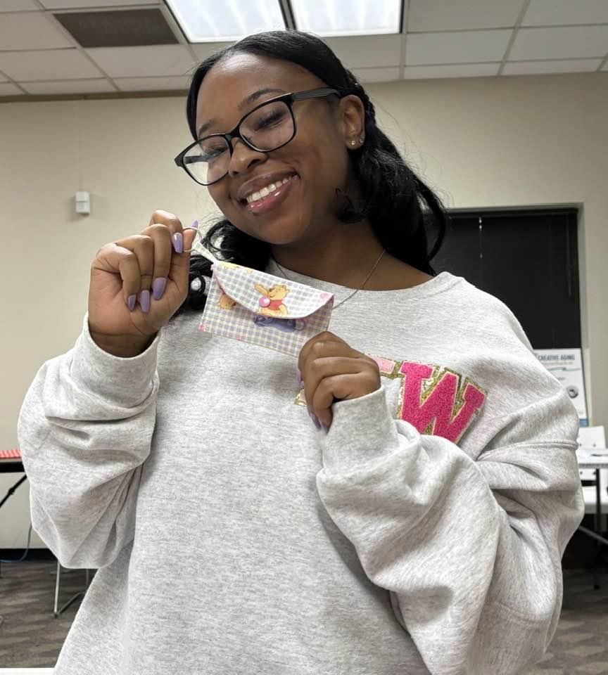 Adult in white sweatshirt smiles and holds up her finished project, a credit card snap wallet.